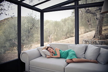 A woman in green dress naps on a sofa in a glass-walled room with olive trees outside. HD wallpaper