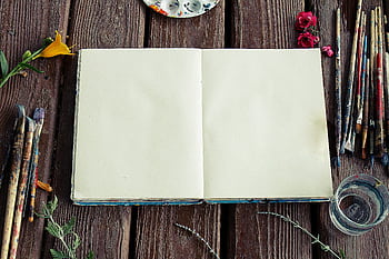 Blank sketchbook surrounded by paintbrushes and flowers on rustic wooden surface. HD wallpaper