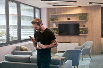 Casual man with tattoos laughing while texting on smart in modern office lounge. HD wallpaper