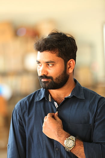 A thoughtful man with a beard and blue shirt posing indoors, displaying a stylish watch. HD phone wallpaper
