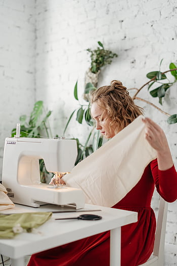 Woman in red dress sewing with a machine in a bright room surrounded by plants. HD phone wallpaper