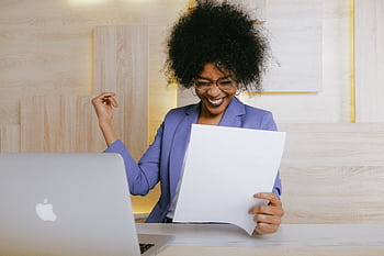 A cheerful woman celebrates her success at work, looking at a document in an office setting. HD wallpaper