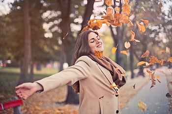 Young woman in a beige coat joyfully throwing autumn leaves in a park. HD wallpaper