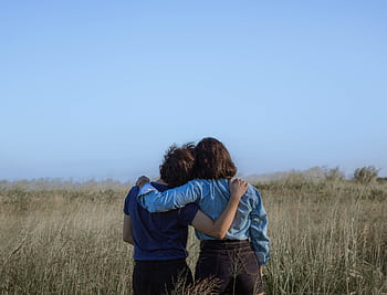 Two individuals share an affectionate embrace in a tranquil grassy field under a clear sky. HD wallpaper