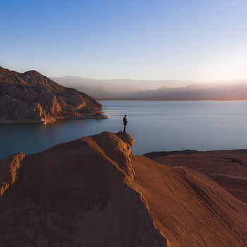 A lone individual stands on a rocky peak overlooking a tranquil lake at sunrise. HD phone wallpaper