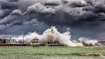 Powerful waves crash against a pier and lighthouse under dark stormy skies. HD wallpaper