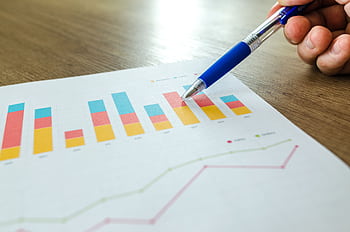 Detailed close-up of a hand pointing at colorful charts with a blue pen on wooden surface. HD wallpaper