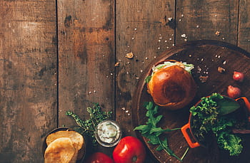 Top view of a rustic wooden table with a burger, vegetables, and condiments, capturing a vintage culinary style. HD wallpaper