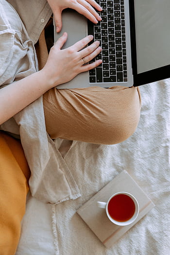 Relaxed home setting featuring a person using a laptop and enjoying tea on a comfortable bed. HD phone wallpaper