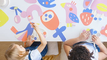 Two children at a table working on colorful paper art cutouts with paintbrushes. HD wallpaper