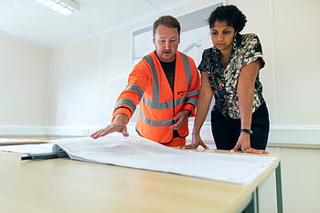 Two engineers reviewing blueprints in an office, discussing project details. HD wallpaper