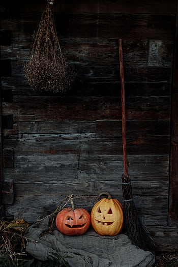 A creepy Halloween scene with carved pumpkins and a witch's broom against a wooden barn wall. HD phone wallpaper