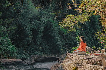 Woman meditating by a serene riverside surrounded by lush greenery on a sunny day. HD wallpaper