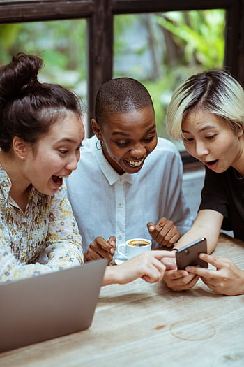 Three friends share a joyful moment browsing a smart together in a cozy café. HD phone wallpaper