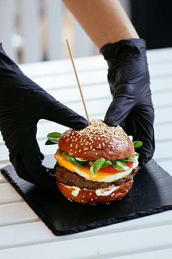 A delicious gourmet burger with sesame bun and fresh vegetables, held by gloved hands on a slate plate. HD phone wallpaper
