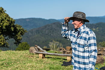 A smiling farmer in plaid flannel enjoys a sunny day on a lush green hillside. HD wallpaper