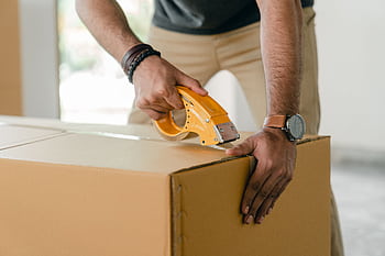 A man sealing a cardboard box using a yellow tape dispenser indoors, focusing on moving or packaging. HD wallpaper