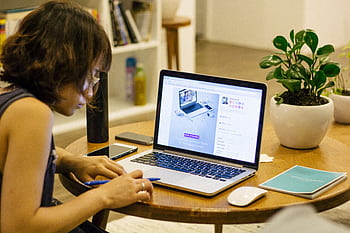 Woman working remotely from home with a laptop, pen, and plant on her desk, highlighting digital nomad lifestyle. HD wallpaper