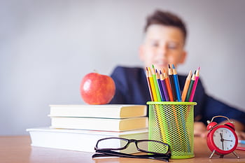Schoolboy smiling behind a desk with books, pencils, and an alarm clock symbolizing study and creativity. HD wallpaper