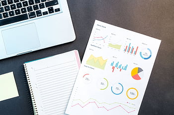 Flatlay of business report with colorful charts, a notebook, and a laptop for data analysis on a desk. HD wallpaper