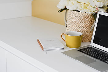 Elegant home workspace setup with a yellow mug, laptop, and flowers in a woven basket on a desk. HD wallpaper