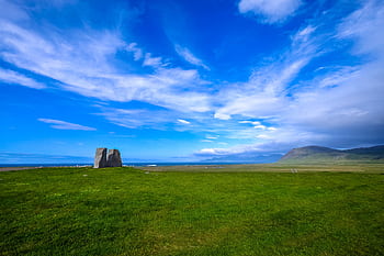 Vast green fields under a vibrant blue sky, featuring distant mountains and a rock formation. HD wallpaper