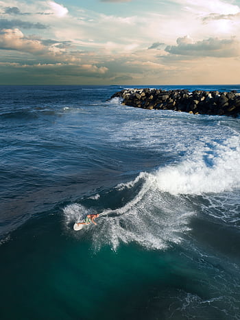 Drone shot capturing a surfer riding waves near a rocky breakwater under a cloudy sky. HD phone wallpaper