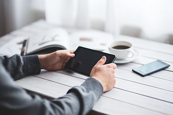 Adult holding a tablet at a minimalist workspace with coffee and a smart nearby. HD wallpaper