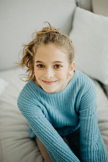 A cheerful young girl with a curly ponytail smiles warmly, wearing a cozy blue knitted sweater. HD phone wallpaper