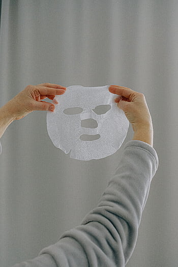 Hands presenting a white sheet mask against a neutral background, promoting skincare and wellness. HD phone wallpaper