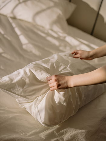 A close-up of hands adjusting a pillow on a neatly made bed, bathed in warm sunlight. HD phone wallpaper