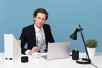A focused businessman in a suit works at his tidy desk with a laptop, plant, and lamp. HD wallpaper