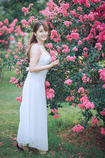 Beautiful woman in a white dress posing by vibrant pink blossoms in a serene park setting. HD phone wallpaper