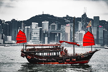 A traditional junk boat with red sails in Hong Kong harbor, against a skyline backdrop. HD wallpaper