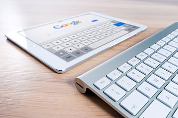A modern tablet displaying a search engine logo next to a wireless keyboard on a wooden desk. HD wallpaper