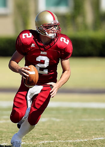Quarterback in red jersey running with football on grassy field during daytime sporting event. HD phone wallpaper