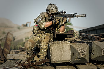 A soldier in full camouflage gear aiming a rifle while in a tactical stance outdoors. HD wallpaper