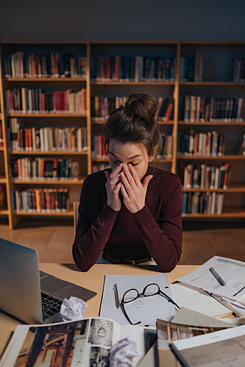 Woman feeling stressed while studying in a library, surrounded by books and laptop. HD phone wallpaper