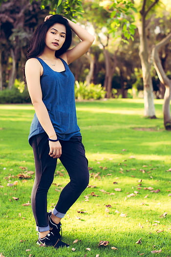 A stylish young woman stands confidently in a lush Dubai park under the sun. HD phone wallpaper