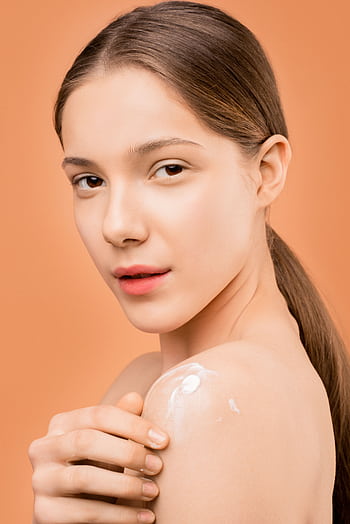 Young woman applies moisturizer on shoulder, set against a peach background, highlighting smooth skin care. HD phone wallpaper