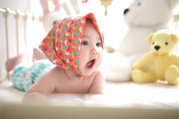 Charming baby with colorful scarf surrounded by cute plush toys, highlighting innocence indoors. HD wallpaper