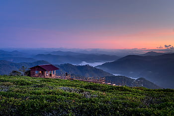 Peaceful mountain landscape at sunrise with a rustic wooden cabin amidst misty hills and lush greenery. HD wallpaper