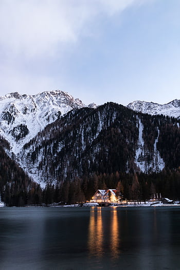 Winter mountain scene with snowy peaks, trees, a tranquil lake reflection, and a cozy cabin. HD phone wallpaper