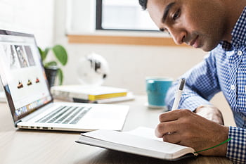 Man focused on writing in a notebook at an office desk with a laptop and coffee cup. HD wallpaper