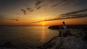 Person sitting on coastal cliffs watching a tranquil sunset over the ocean, creating a peaceful and scenic view. HD wallpaper