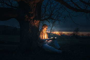 A man reading under a tree at night, illuminated by a glowing book. HD wallpaper