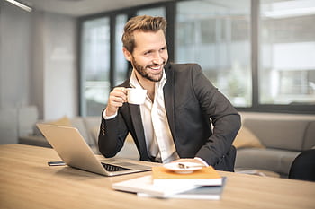 Smiling businessman in formal attire enjoying coffee break at modern office desk with laptop. HD wallpaper