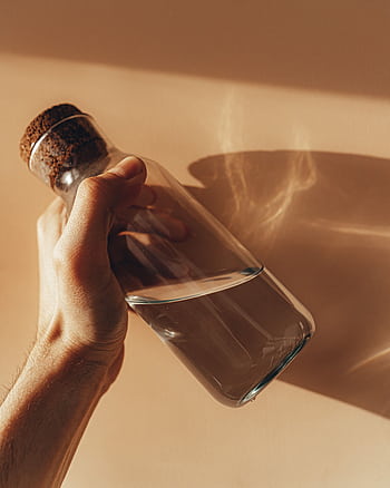 Close-up of a hand holding a clear glass bottle with water, casting a shadow on a beige wall. HD phone wallpaper
