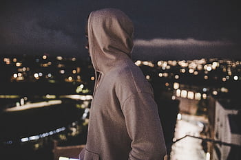 A person in a hoodie overlooks a blurry cityscape at night, evoking mystery and solitude. HD wallpaper