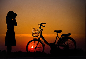 A silhouette of a woman grapher and a bicycle against a vibrant sunset background. HD wallpaper